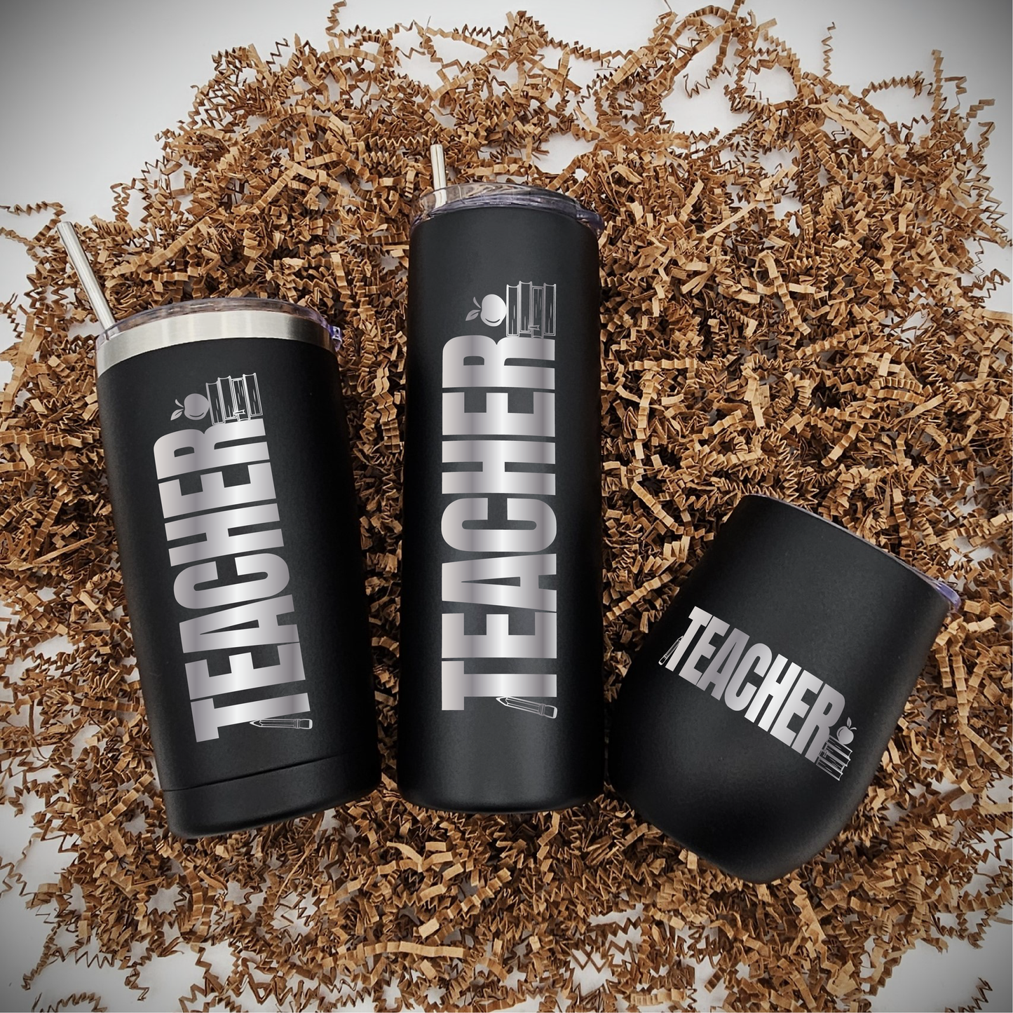 Three tumblers of varying sizes, each with 'teacher, a set of books, an apple, and a pencil' laser-engraved on the front, placed on brown crinkled paper.