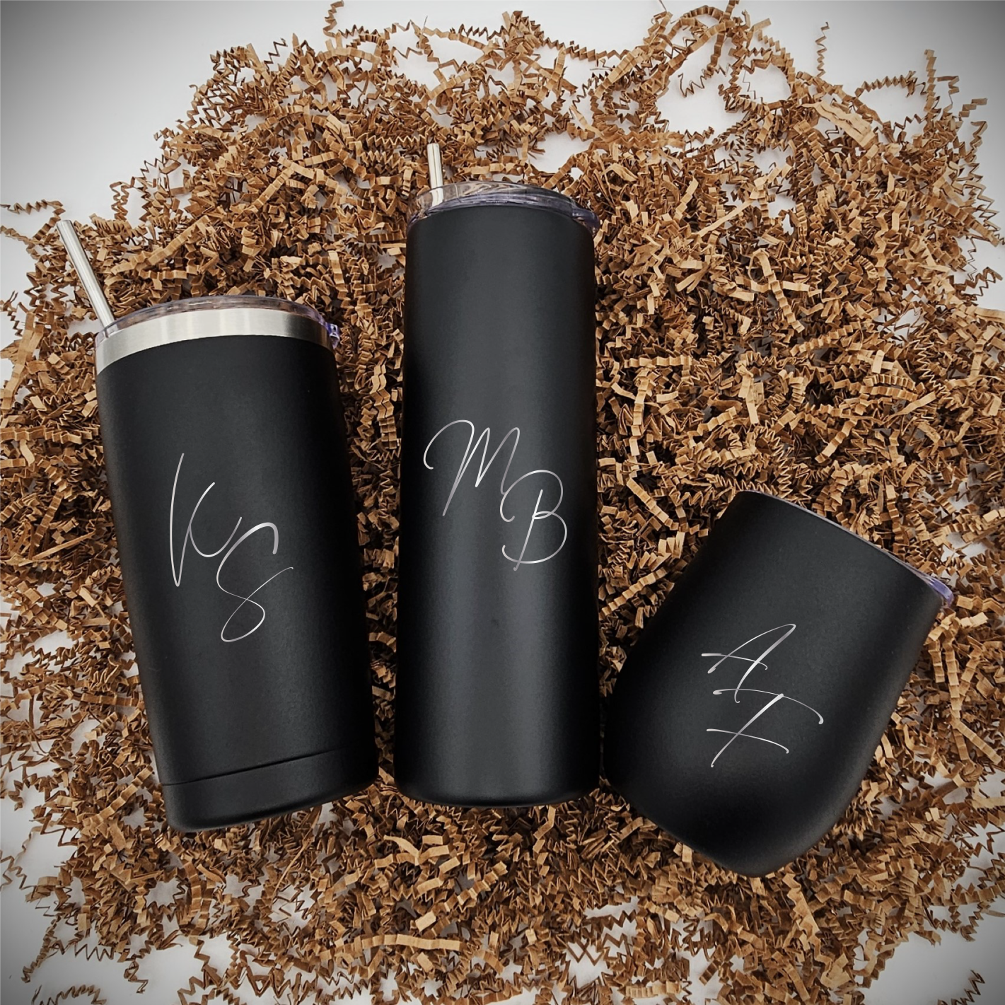 Three stainless steel tumblers of various sizes with laser engraved initials facing forward, arranged on crinkled brown paper.