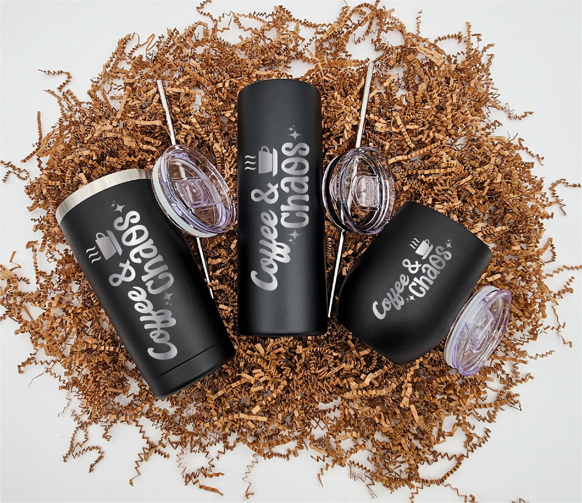 Three stainless steel tumblers of varying sizes, each engraved with 'Coffee & Chaos' vertically, resting on crinkled brown paper.