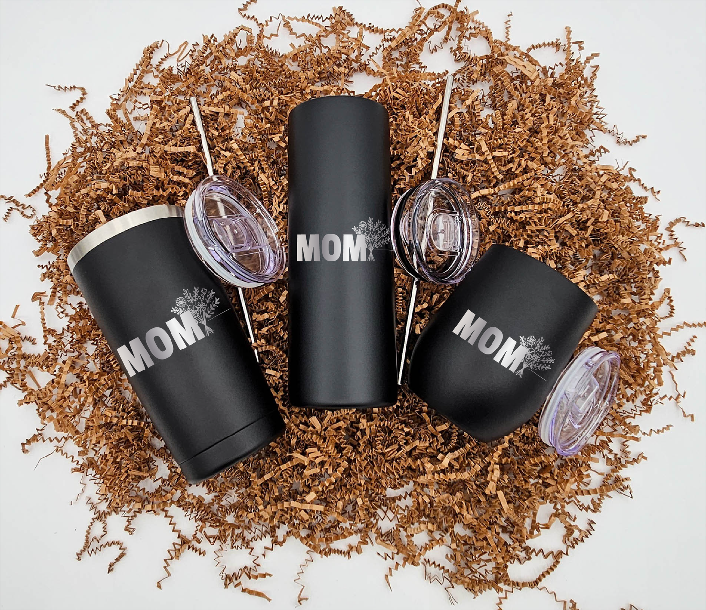 Three stainless steel tumblers of different sizes, each engraved with 'Mom' and a bouquet of flowers horizontally, positioned on crinkled brown paper.