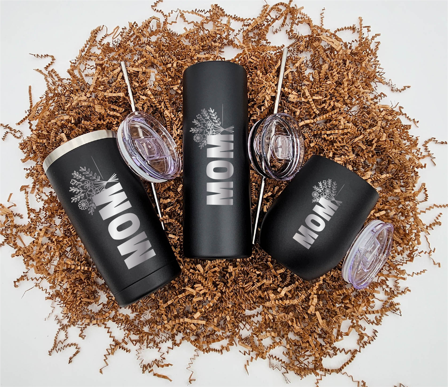 Three stainless steel tumblers of different sizes, each engraved with 'Mom' and a bouquet of flowers vertically, positioned on crinkled brown paper.