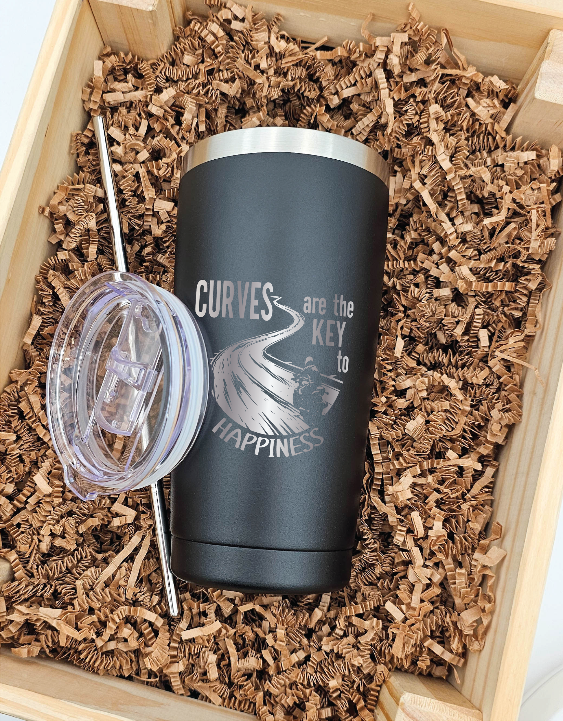 20 ounce stainless steel tumbler featuring a laser-engraved image of a motorcycle cruising along a winding road, accompanied by the text 'curves are the key to happiness'. The tumbler is displayed forward-facing on crinkled brown paper, with its lid and straw placed beside it.