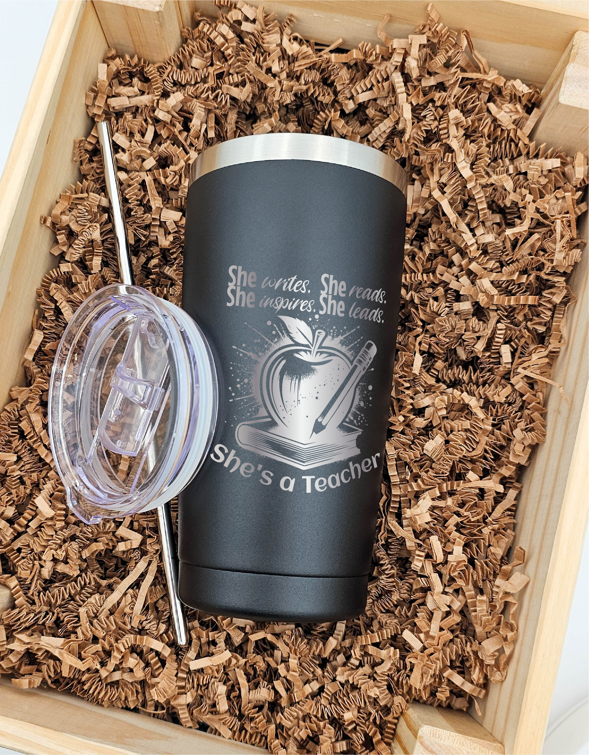 Front view of a 20 oz. tumbler against a brown crinkled paper backdrop, featuring laser engraving of a book, apple, and pencil. Engraved text reads: 'She writes, she reads, she inspires, she leads; she's a teacher.