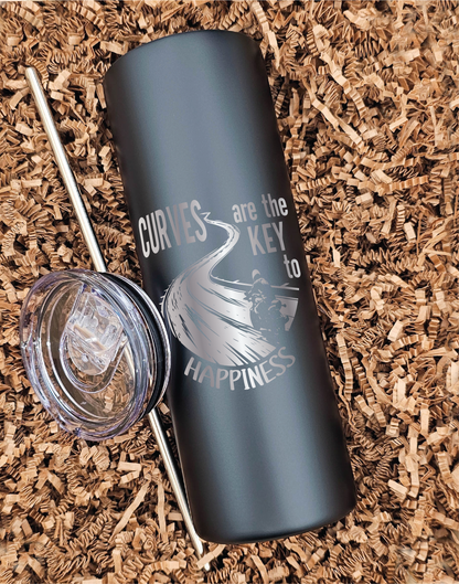 20 oz skinny laser-engraved motorcycle tumbler with lid and straw beside it, featuring a motorcycle on a curvy road with the phrase 'Curves are the key to happiness', displayed on brown crinkled paper.