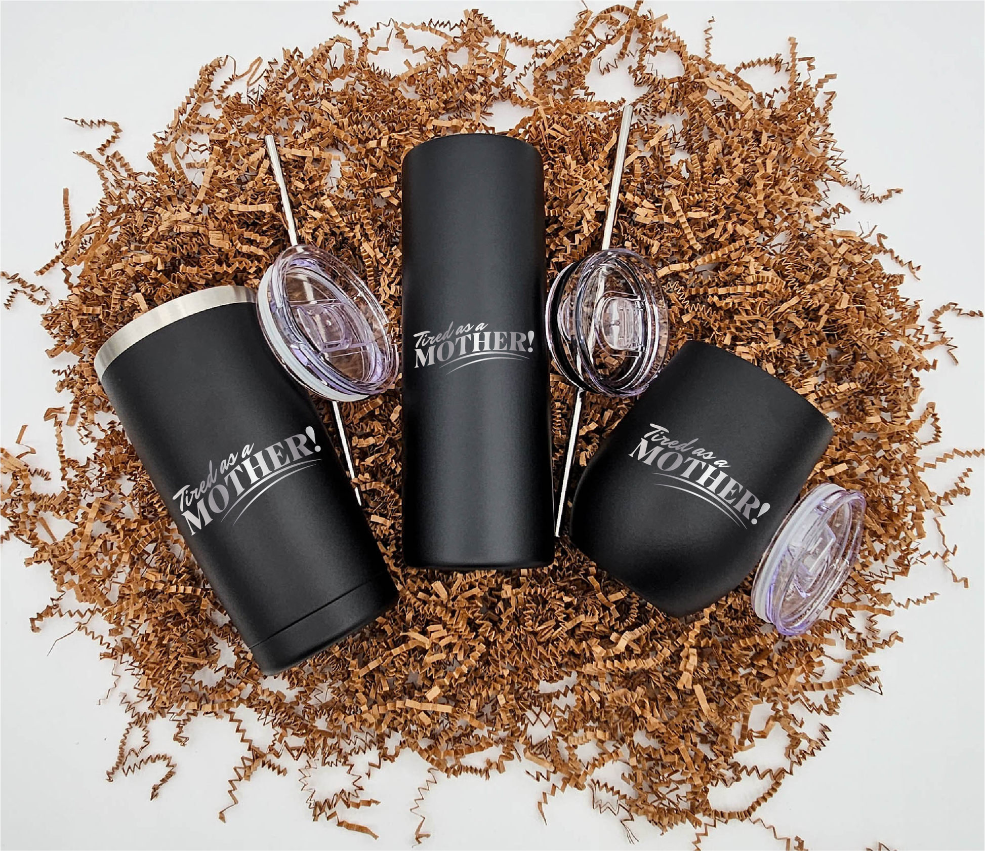 Three stainless steel tumblers of varying sizes, each engraved with 'Tired as a Mother' horizontally, resting on crinkled brown paper.