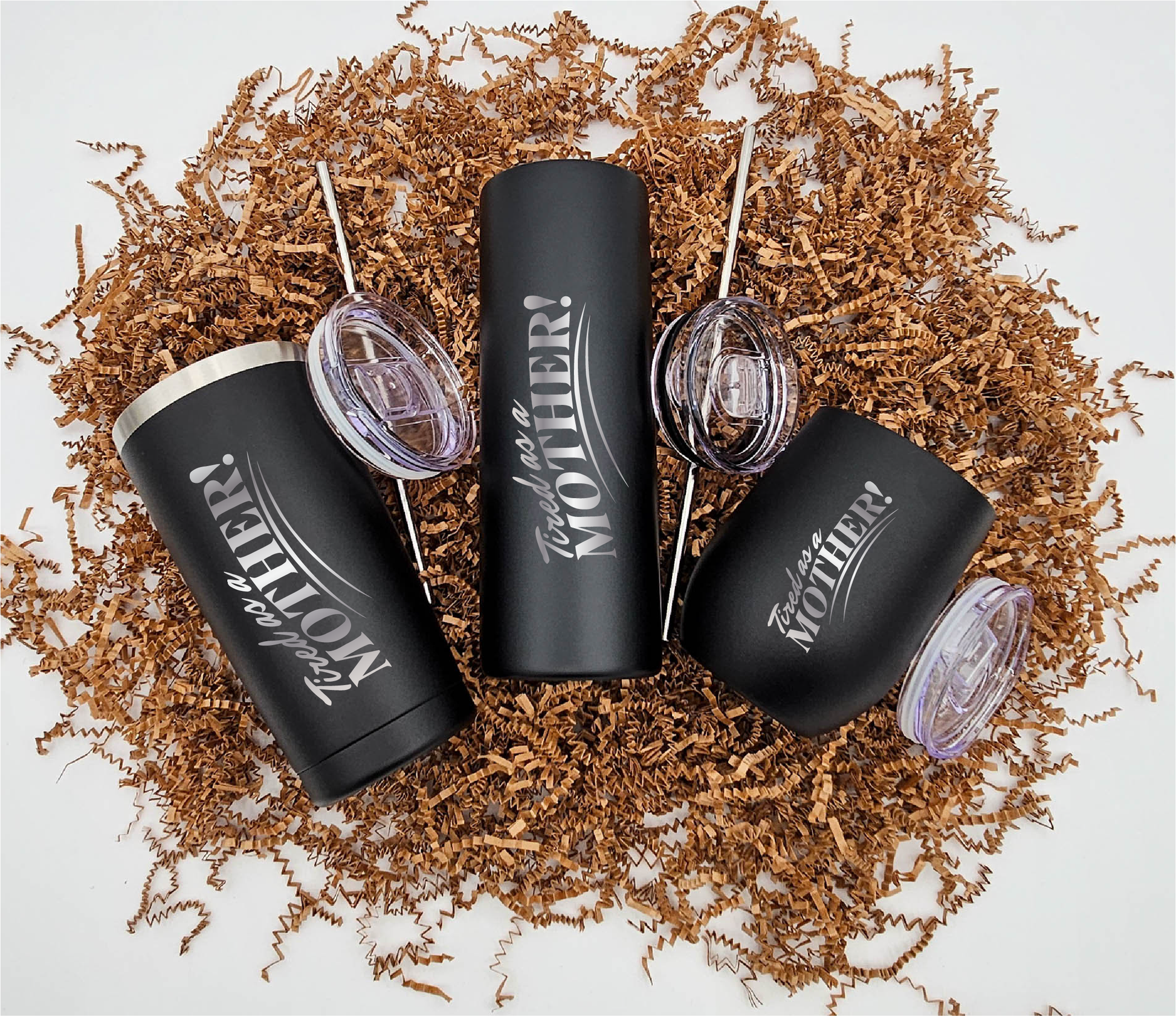 Three stainless steel tumblers of varying sizes, each engraved with 'Tired as a Mother' vertically, resting on crinkled brown paper.