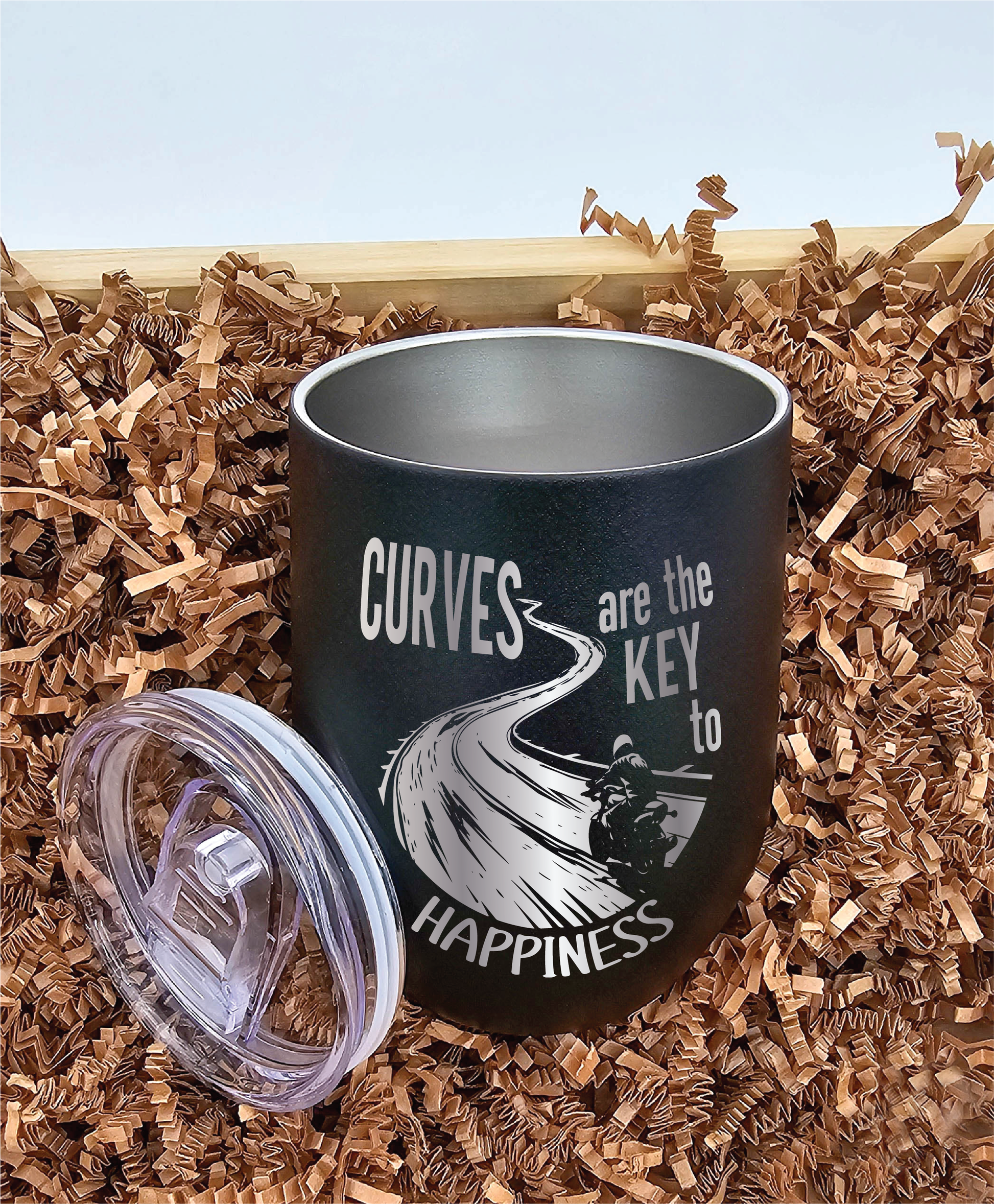 12 oz stainless steel tumbler featuring laser-engraved motorcycle on a winding road with the inscription 'Curves are the key to happiness.' Lid shown, resting on brown crinkled paper.