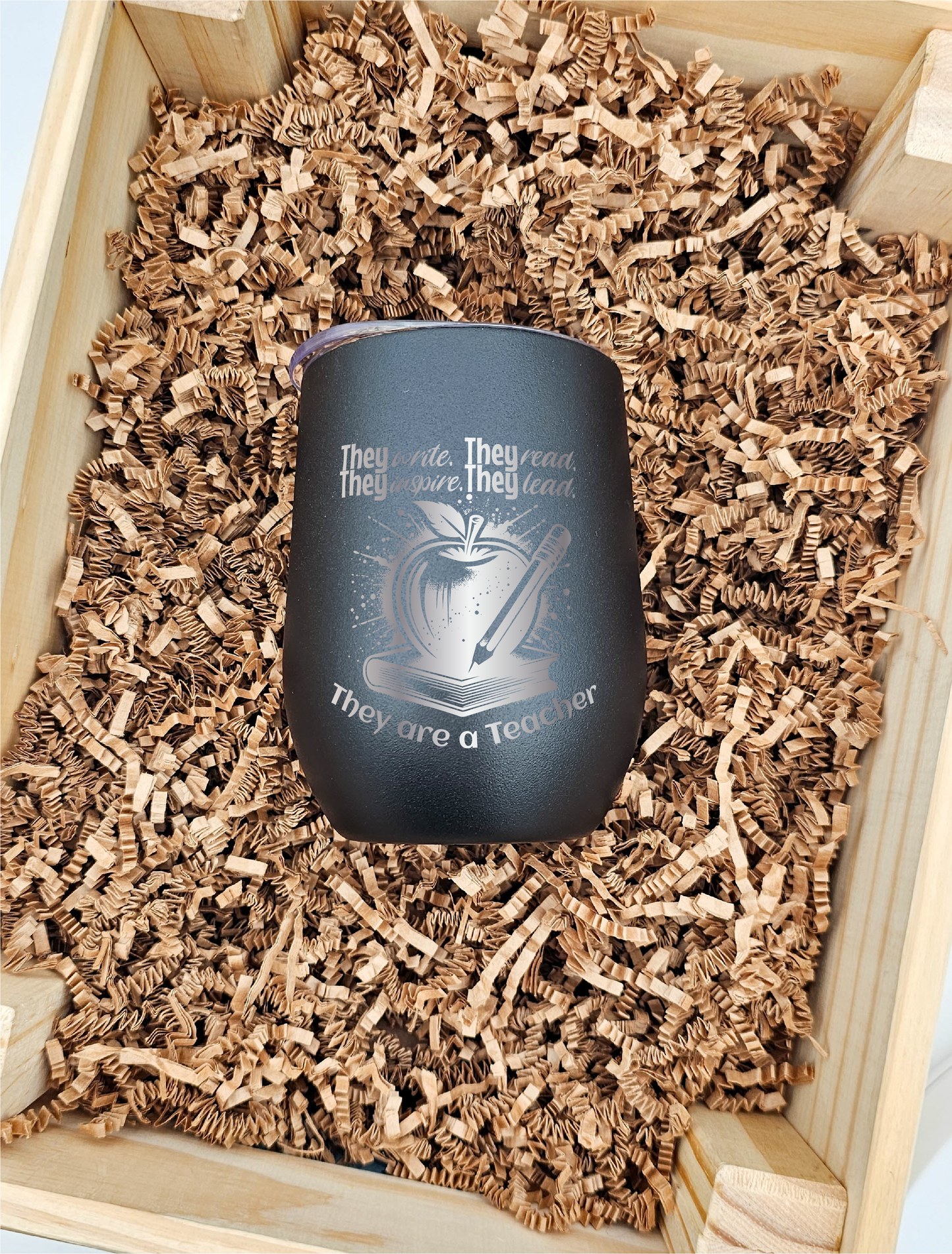 Front-facing view of a 12 oz. wine tumbler against a textured brown crinkled paper backdrop. The tumbler is laser engraved with symbols of education – a book, apple, and pencil – alongside the phrase 'They write, they read, they inspire, they lead; they are a teacher.' Perfect for honoring the dedication of educators.